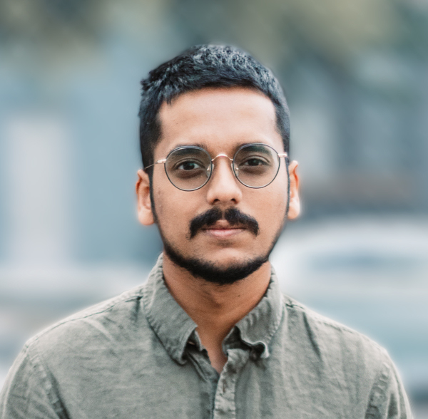 Portrait of a person with short hair, and dark tinted glasses, they have facial hair and are wearing an olive green button down shirt, the background is portrayed in shallow depth of field blurred effect.