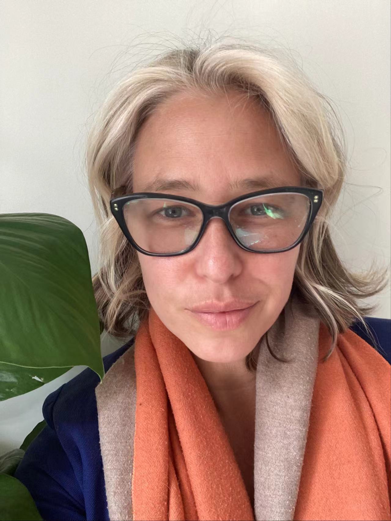 portrait of maya kramer, wearing black glasses and an orange scarf. in the background there is a single green leaf peaking over her shoulder
