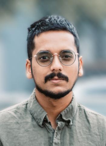 Portrait of a person with short hair, and dark tinted glasses, they have facial hair and are wearing an olive green button down shirt, the background is portrayed in shallow depth of field blurred effect.