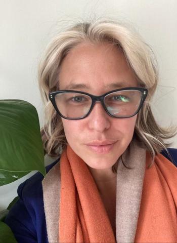 portrait of maya kramer, wearing black glasses and an orange scarf. in the background there is a single green leaf peaking over her shoulder